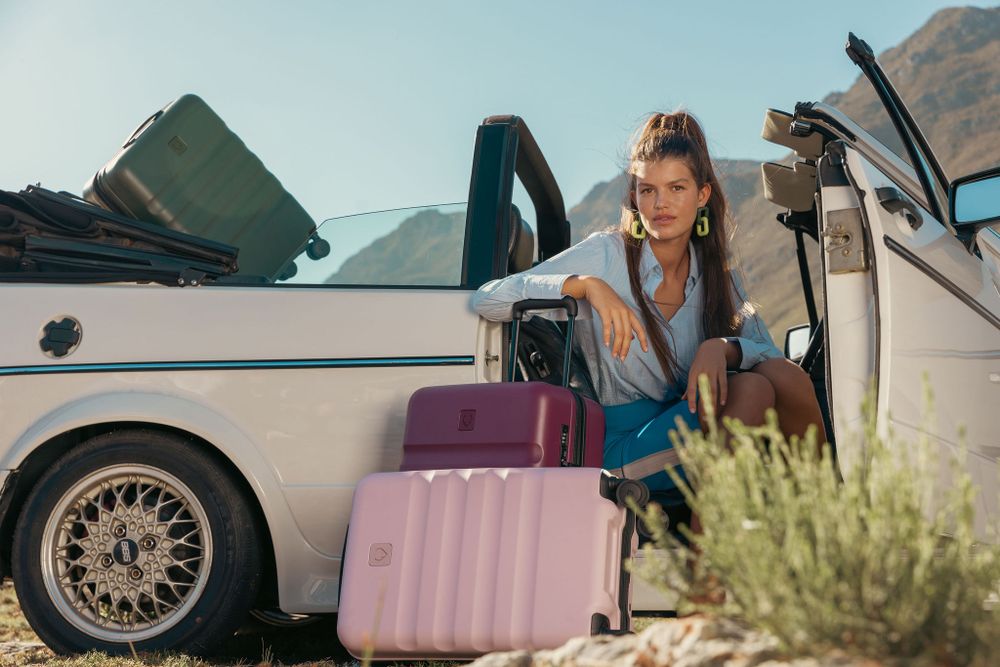 Woman in blue outfit loading pink luggage into vintage car with mountains in background.