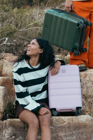 Person in striped sweater sitting with pink and green luggage against rocky backdrop.