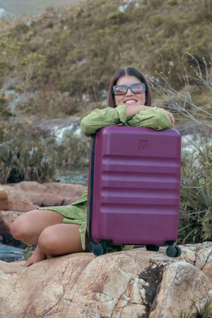 Person resting on purple rolling suitcase against rocky mountain landscape.