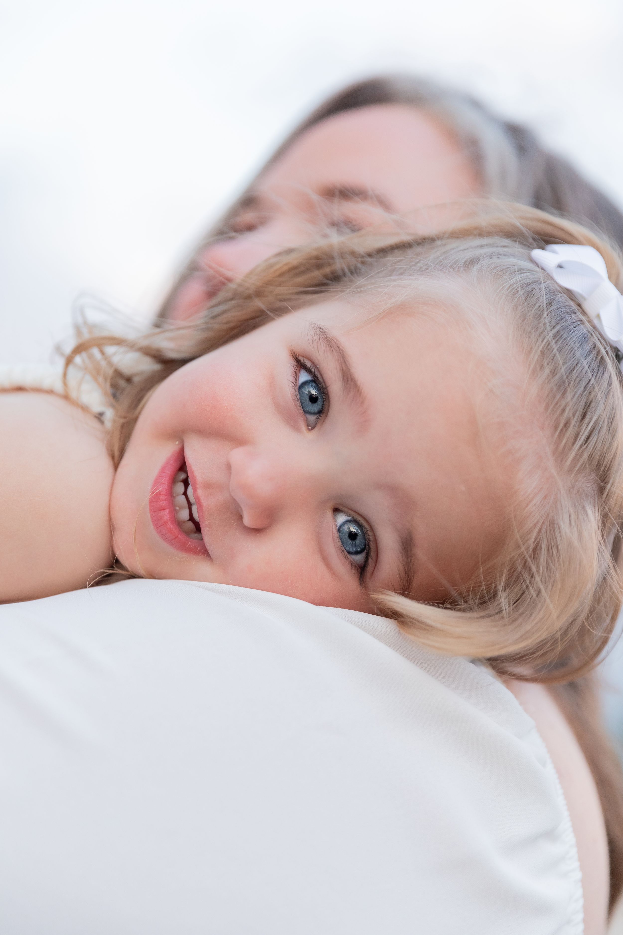 Young child with bright blue eyes and blonde hair lies on white bedding with a joyful smile.