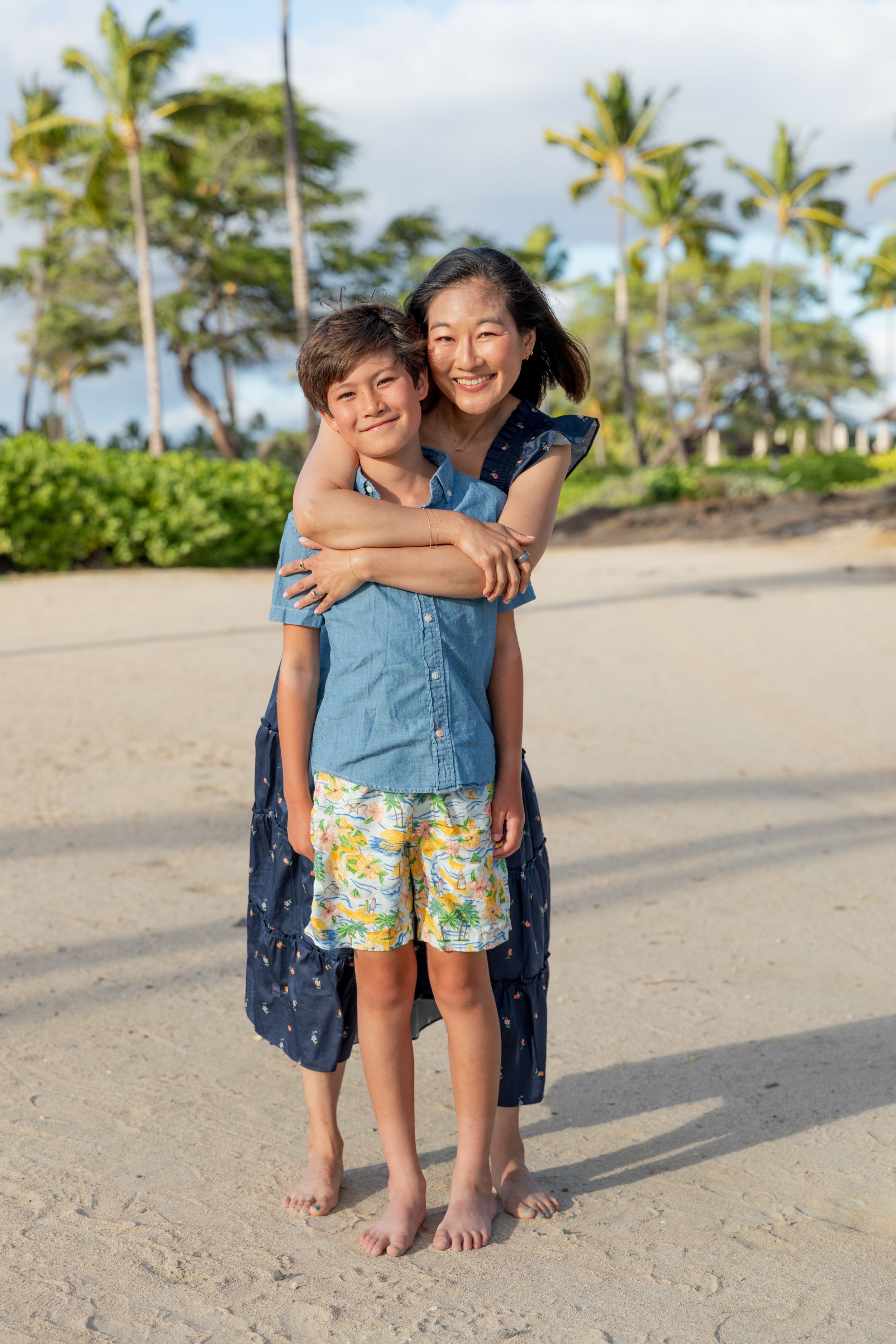 Two people share a warm embrace on a tropical beach with palm trees and sand in the background.