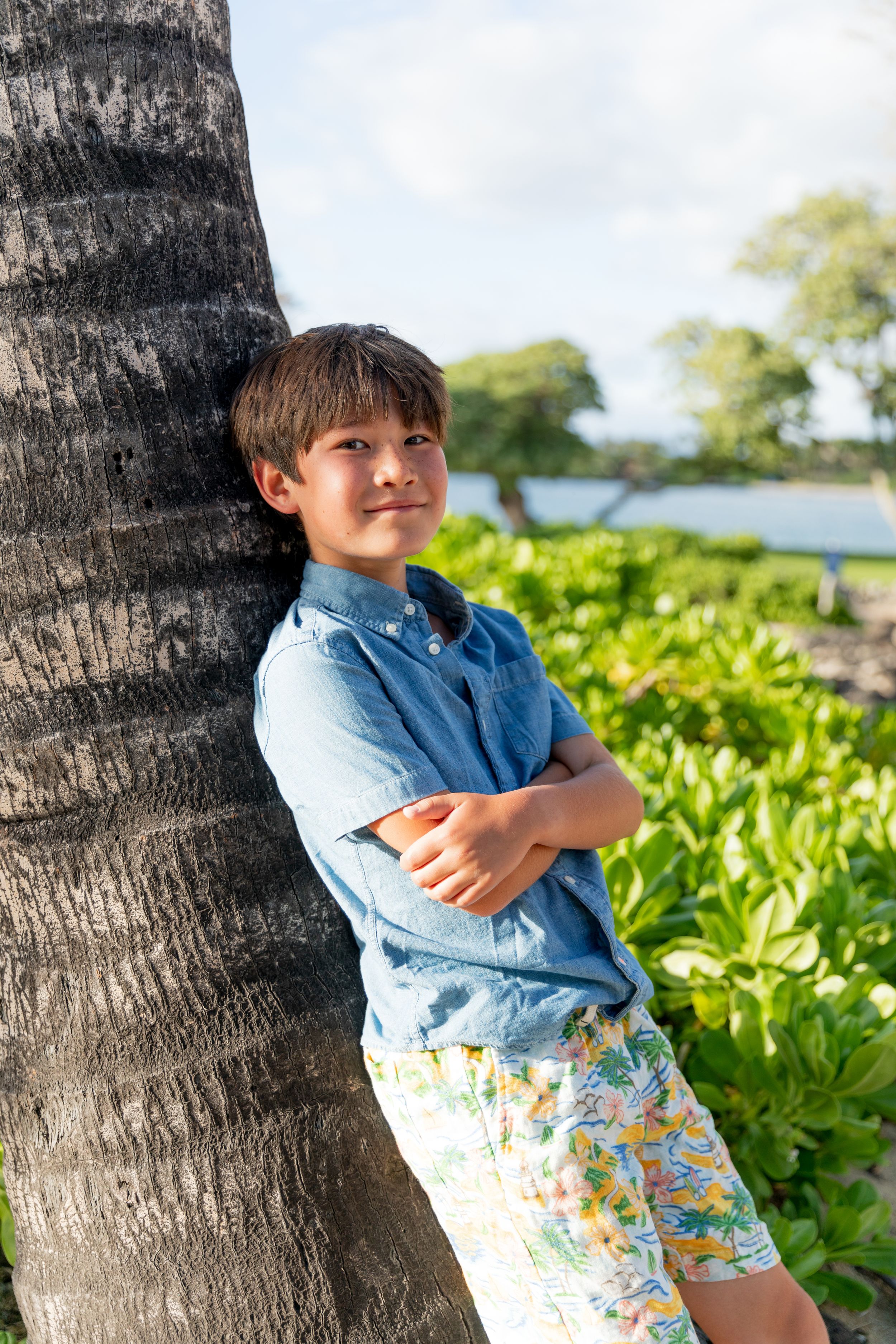 A young person in a blue shirt and floral shorts leans against a palm tree in a tropical setting.