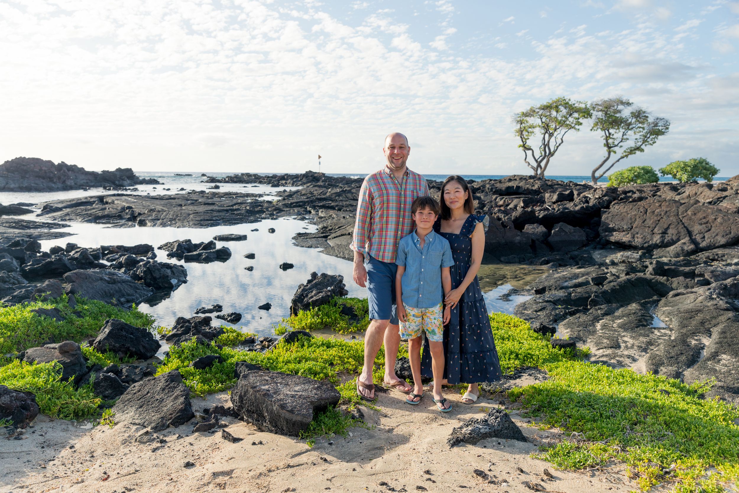 Three people stand together on volcanic rocks by the ocean with greenery and blue sky in Hawaii.