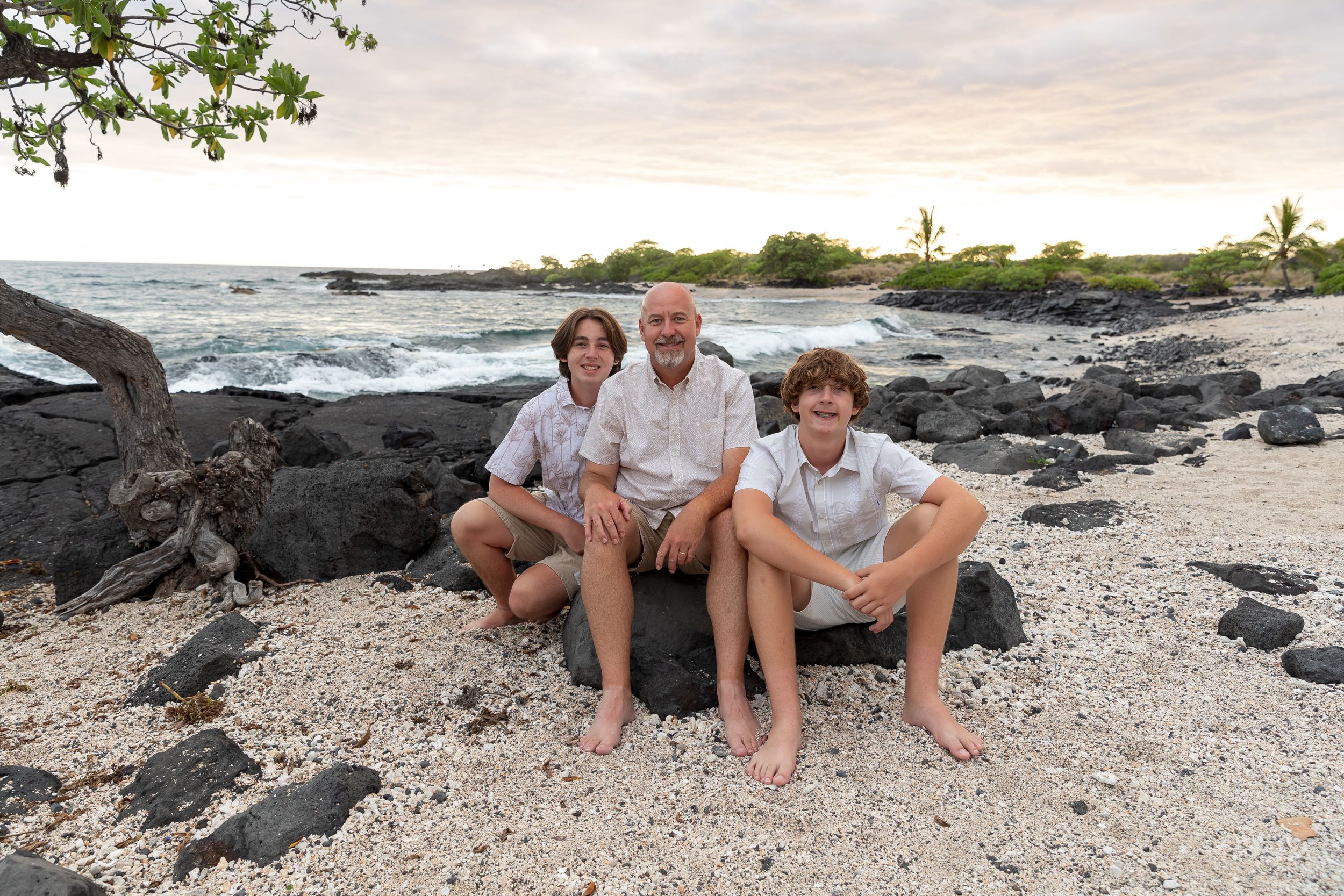 Three people sit together on a rocky beach with ocean waves and palm trees in the background.