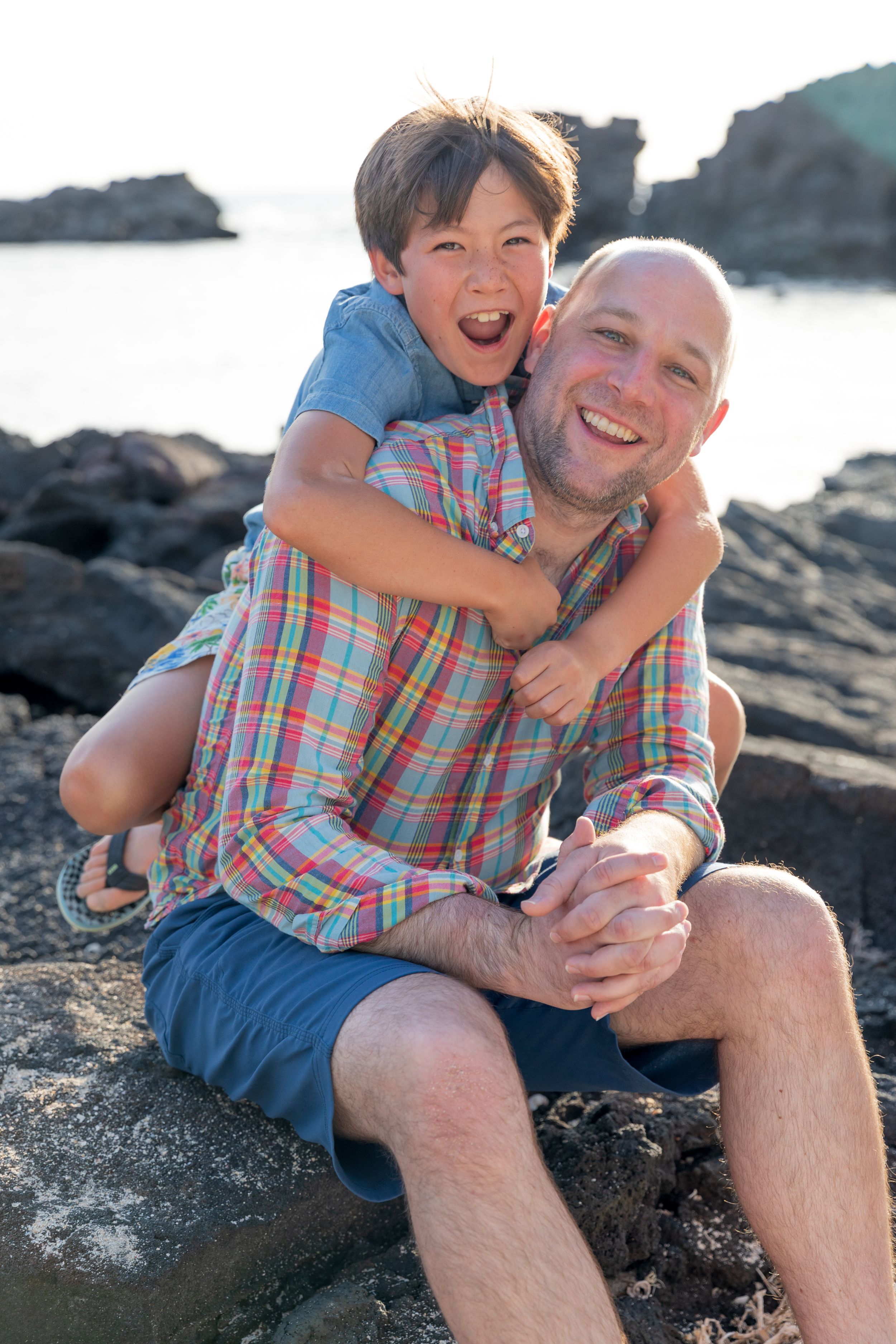 Two people share a joyful moment on black volcanic rocks by the ocean at sunset.