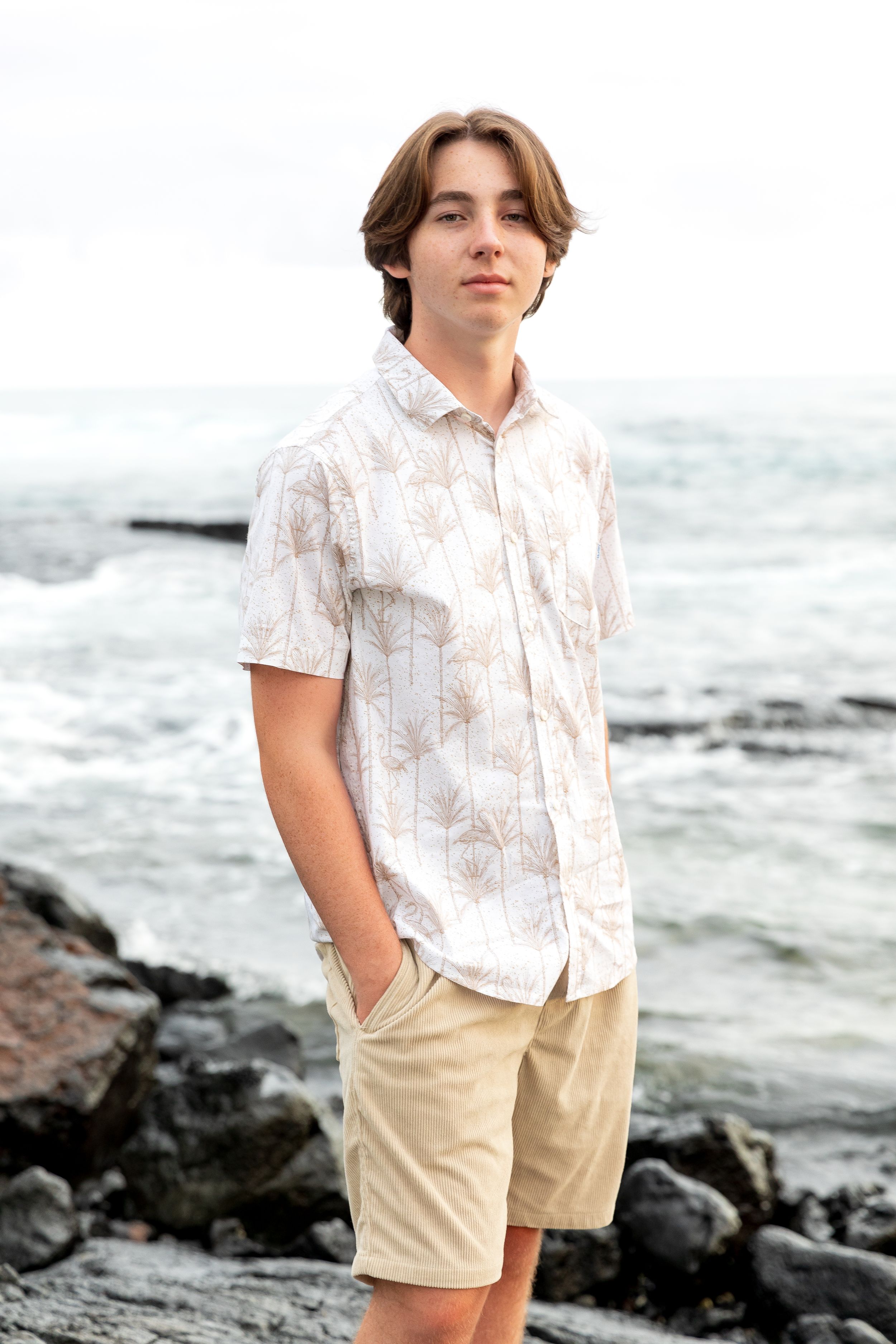 A person in a patterned white shirt and khaki shorts poses against a rocky coastal backdrop.
