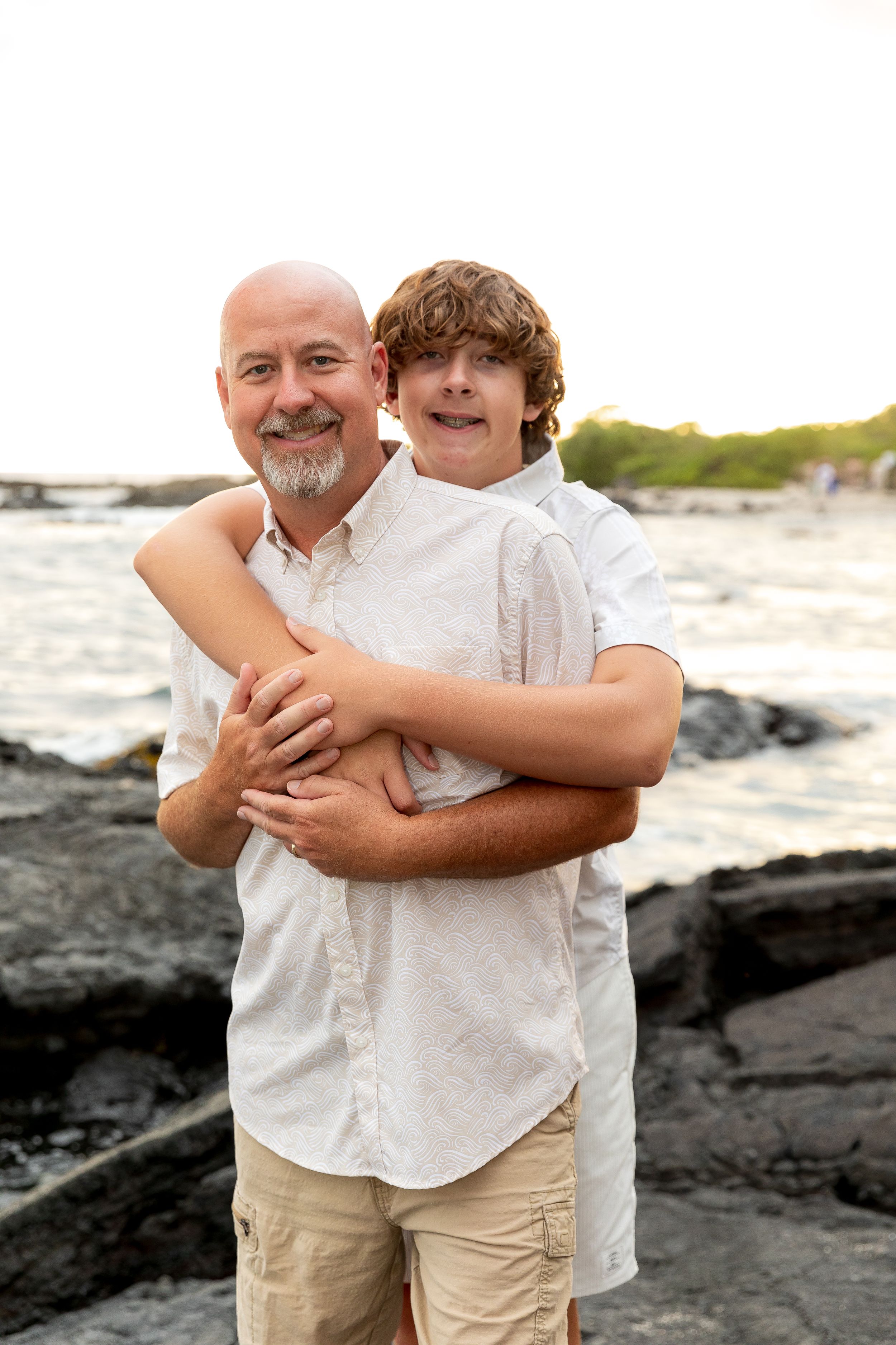 Two people share an embrace while standing on black volcanic rocks near the ocean.