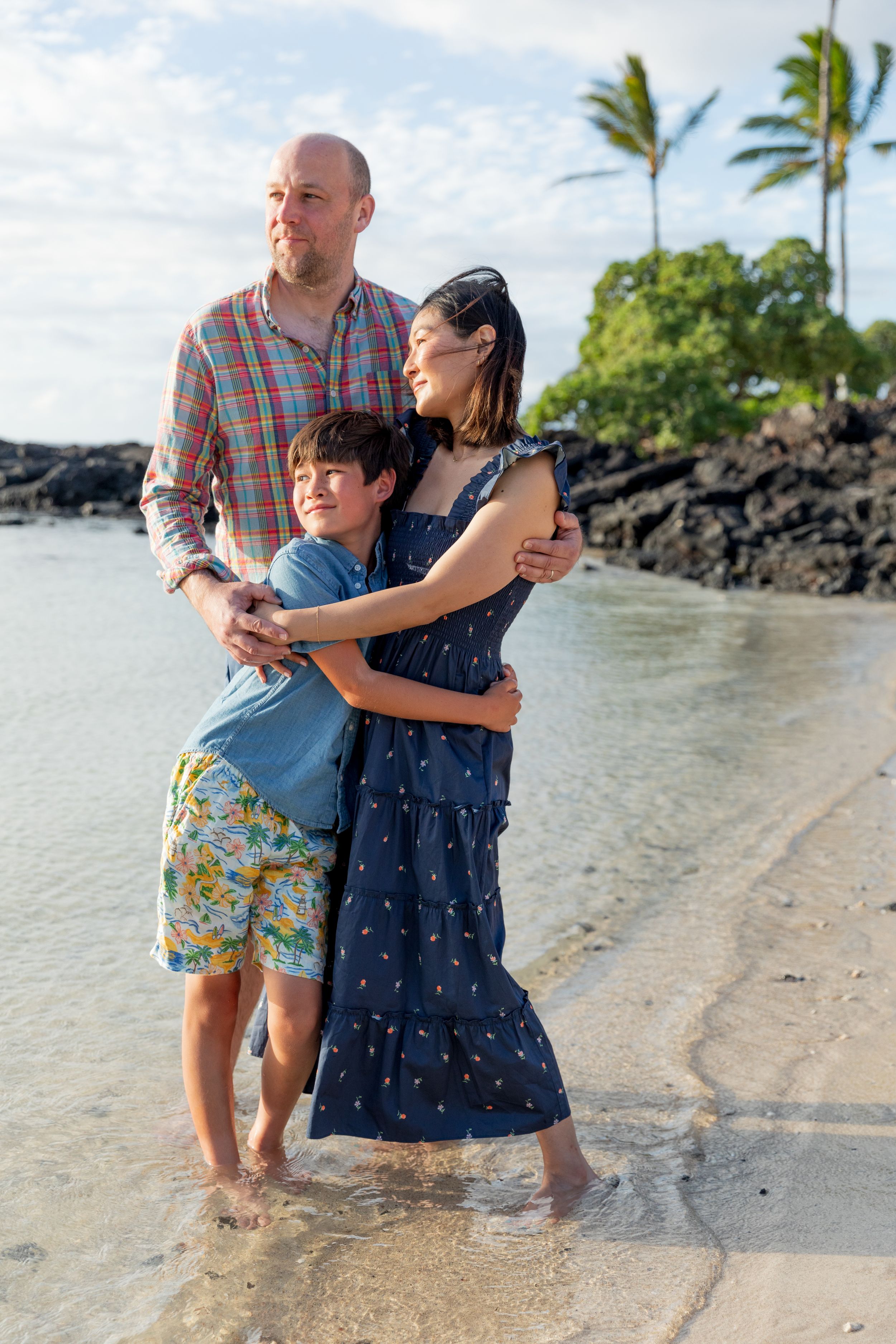 Three people stand together in shallow ocean water on a tropical beach with palm trees and volcanic rocks.