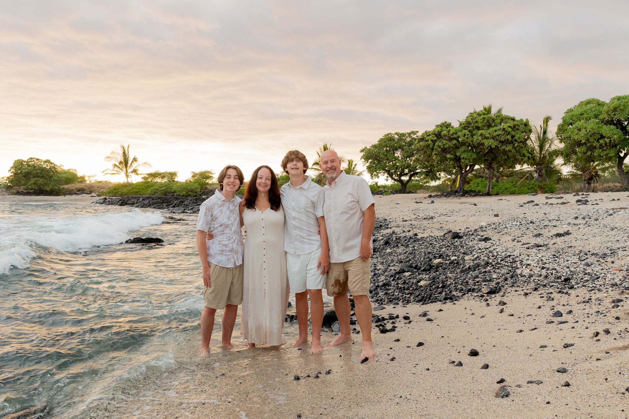 A family group poses together at sunset on a Hawaiian beach with volcanic rocks and palm trees.