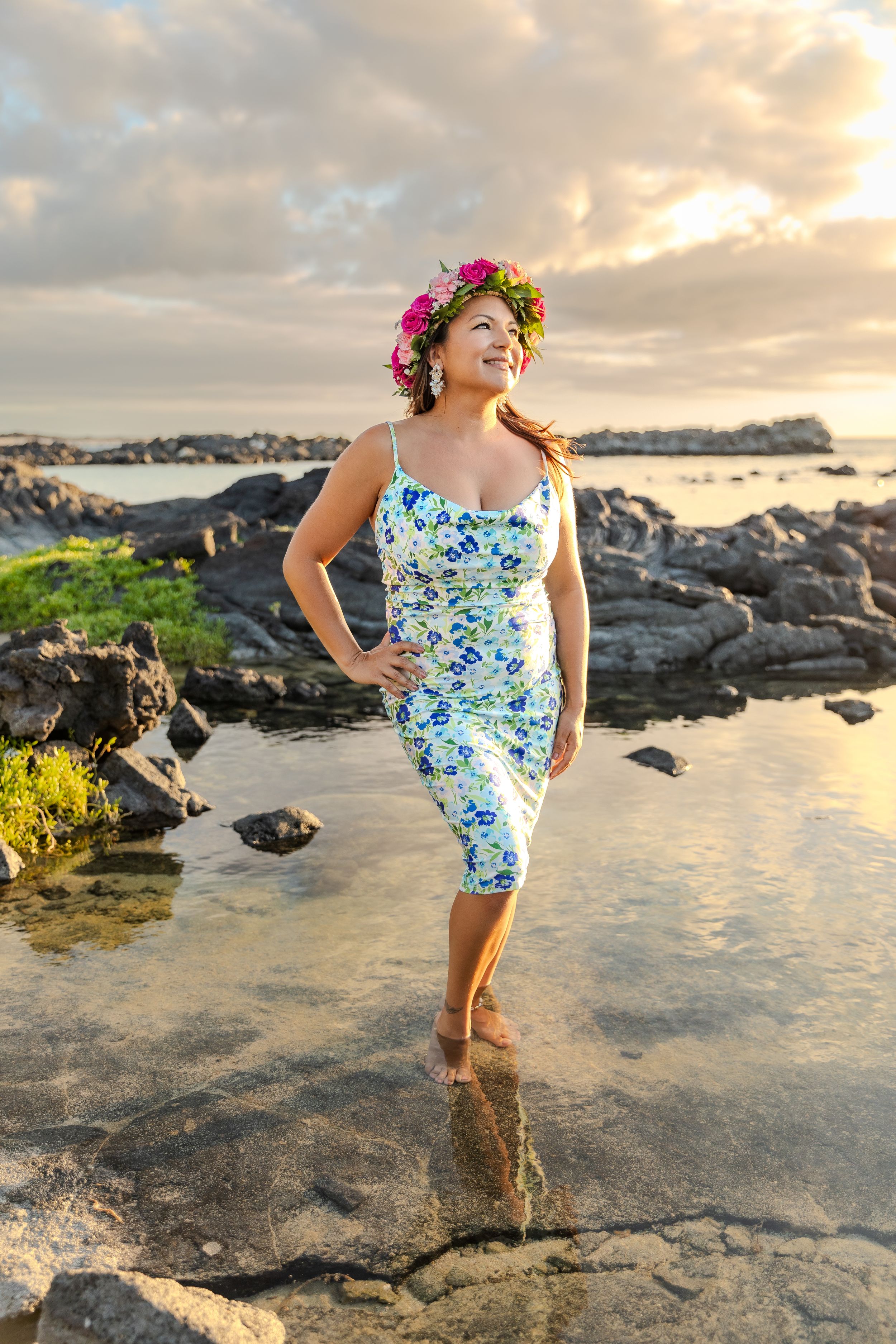 A woman in a floral dress and flower crown poses on volcanic rocks at sunset.