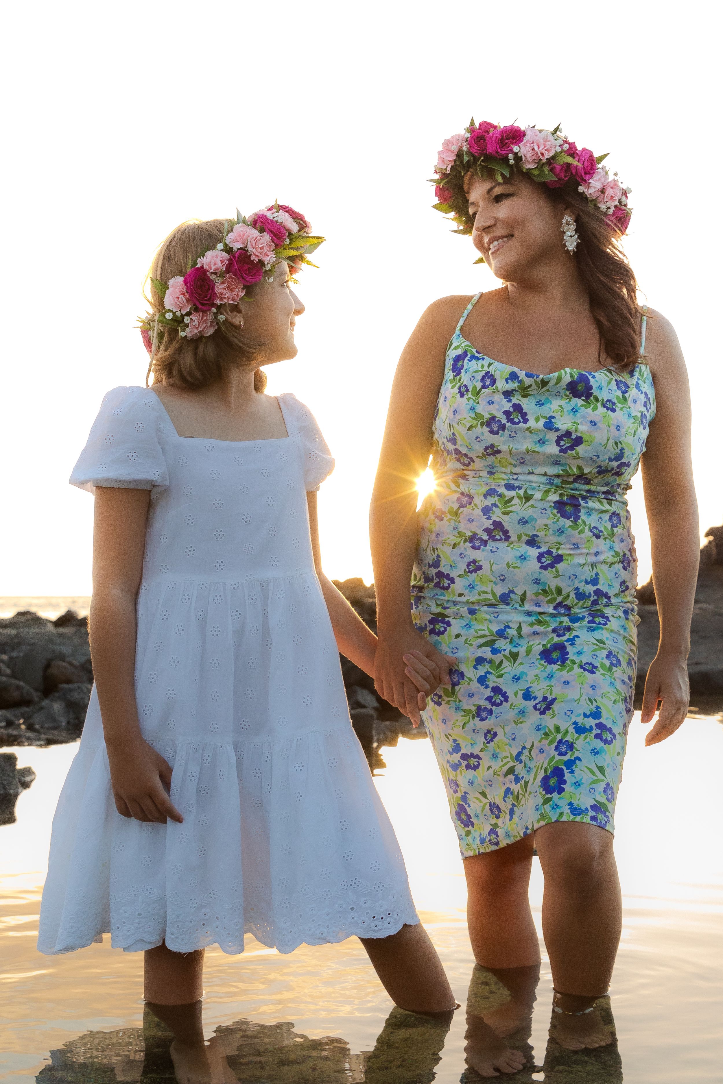 Two people in summer dresses and flower crowns stand on rocky shore at sunset.