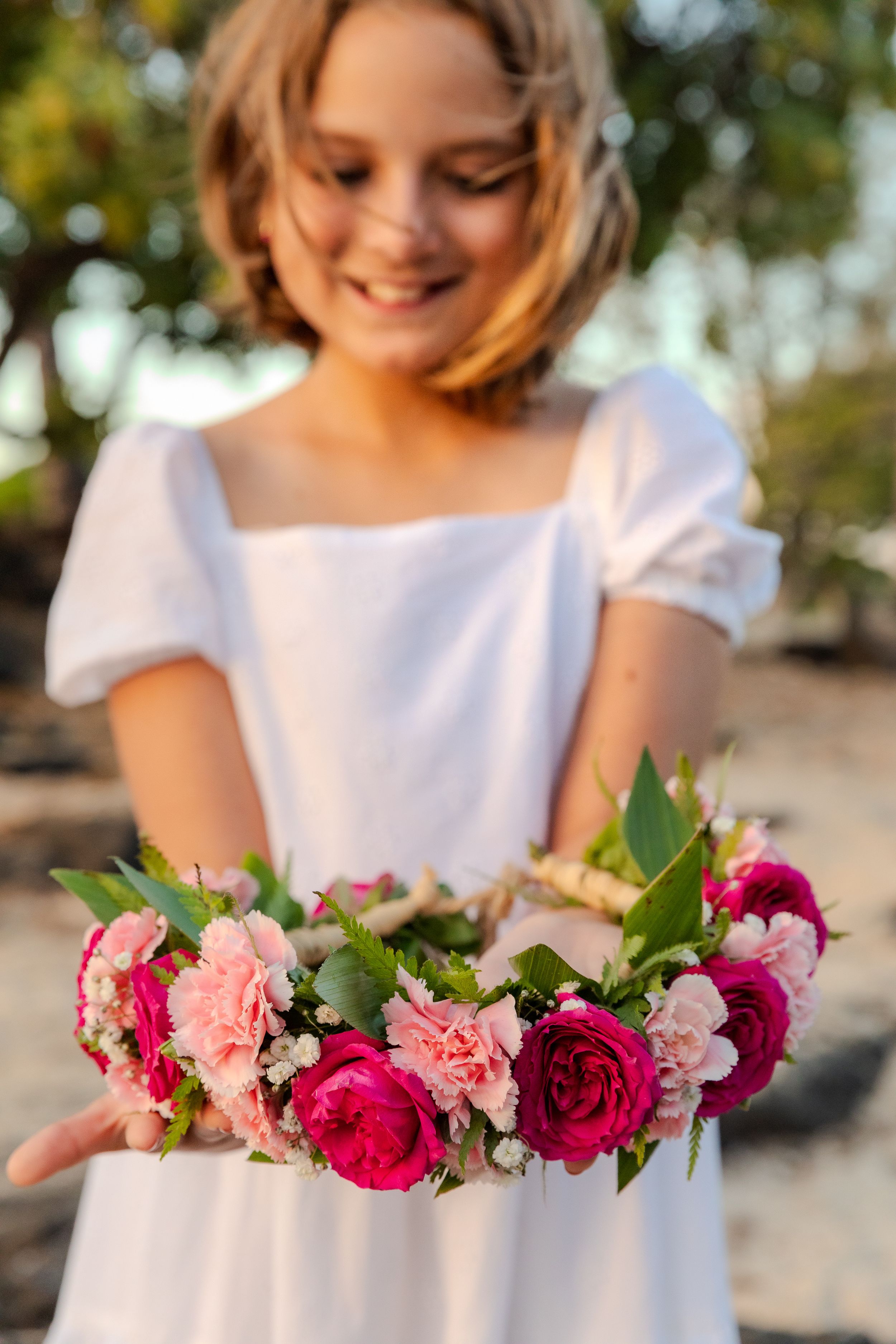 A pink and red flower crown held against a white dress in warm sunlight.