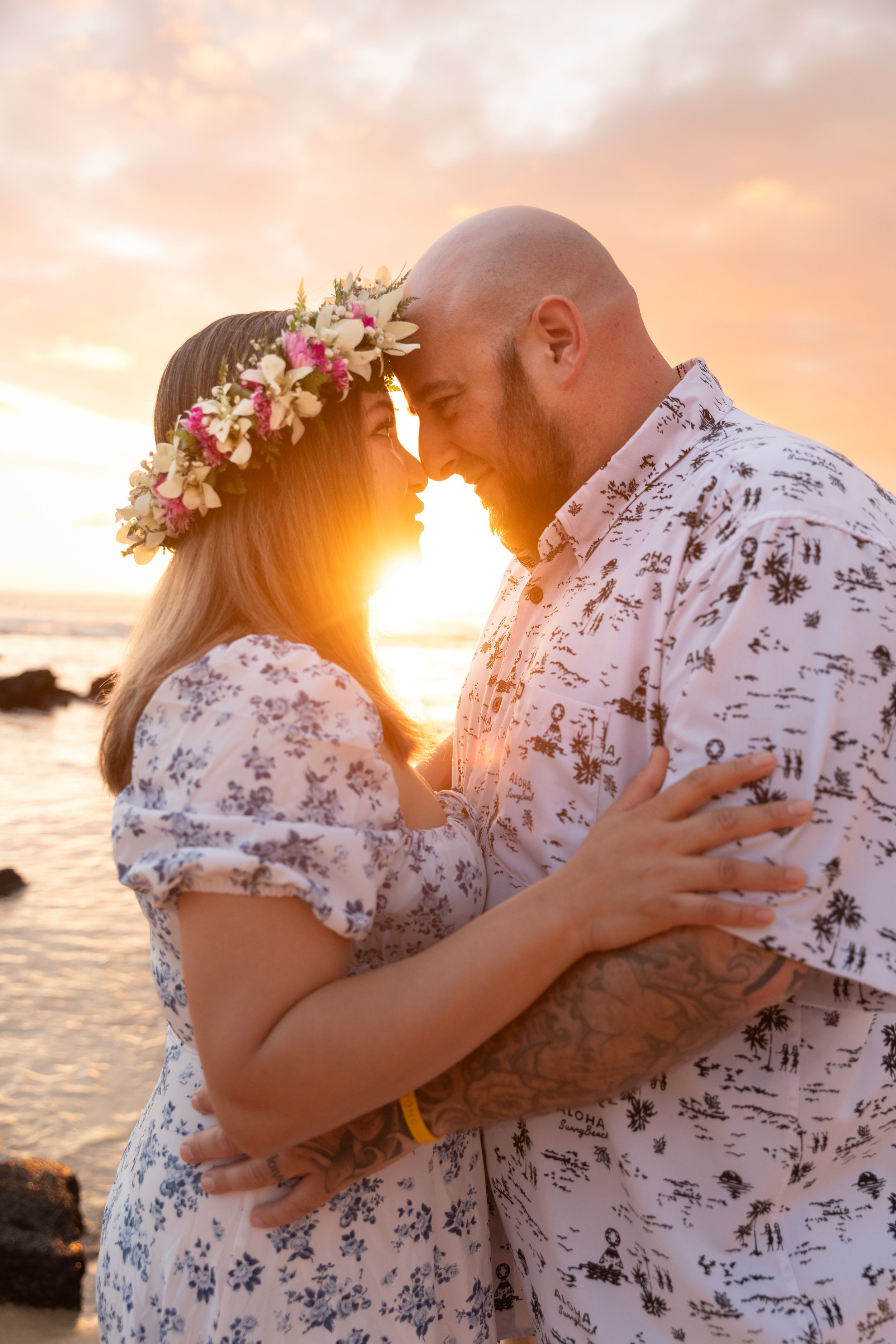 Romantic couple in matching floral outfits embrace during golden hour sunset on a rocky beach.