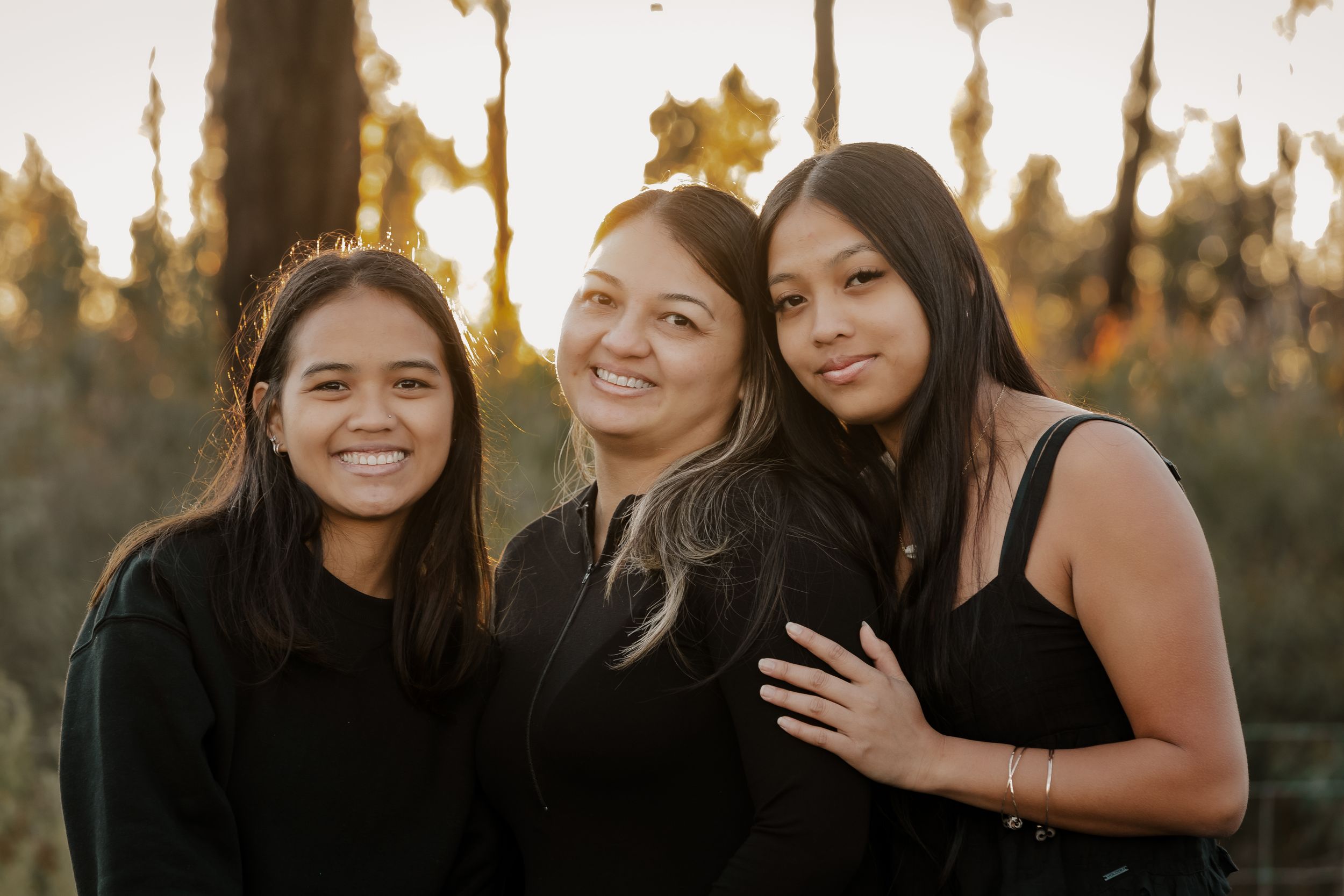 Three friends smile together during golden hour with trees in the background.