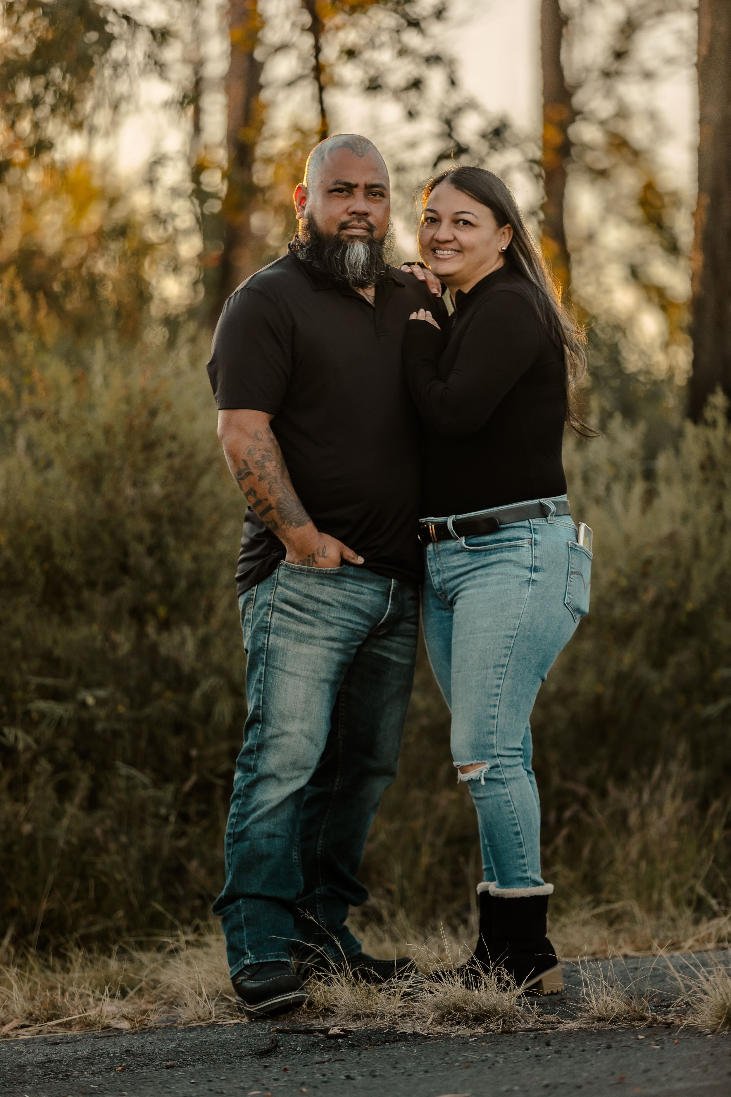 A couple poses together outdoors at sunset among tall trees.