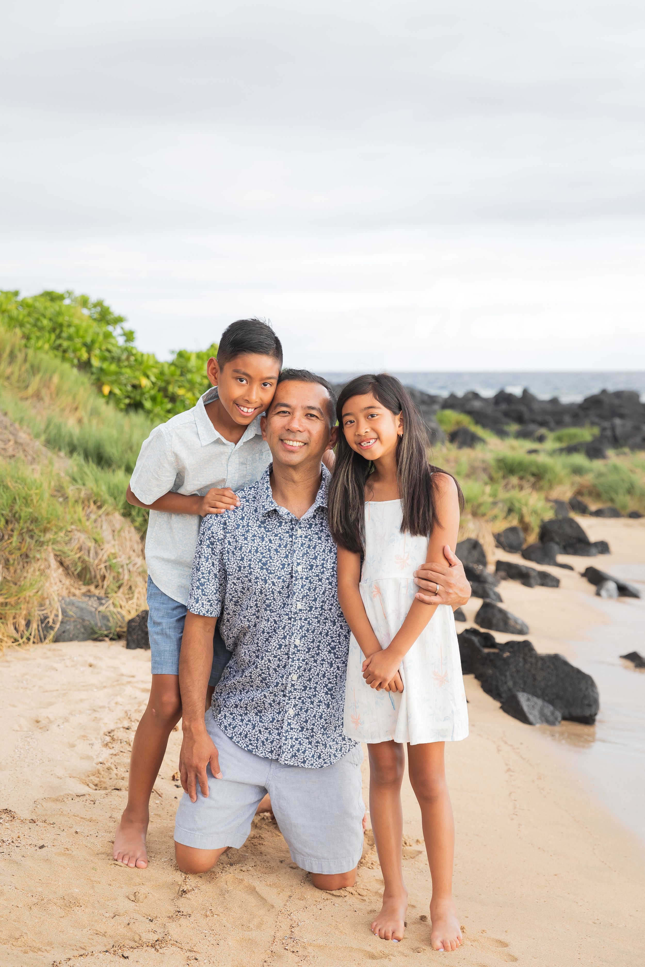 Three people pose together on a sandy beach path with volcanic rocks and ocean views.