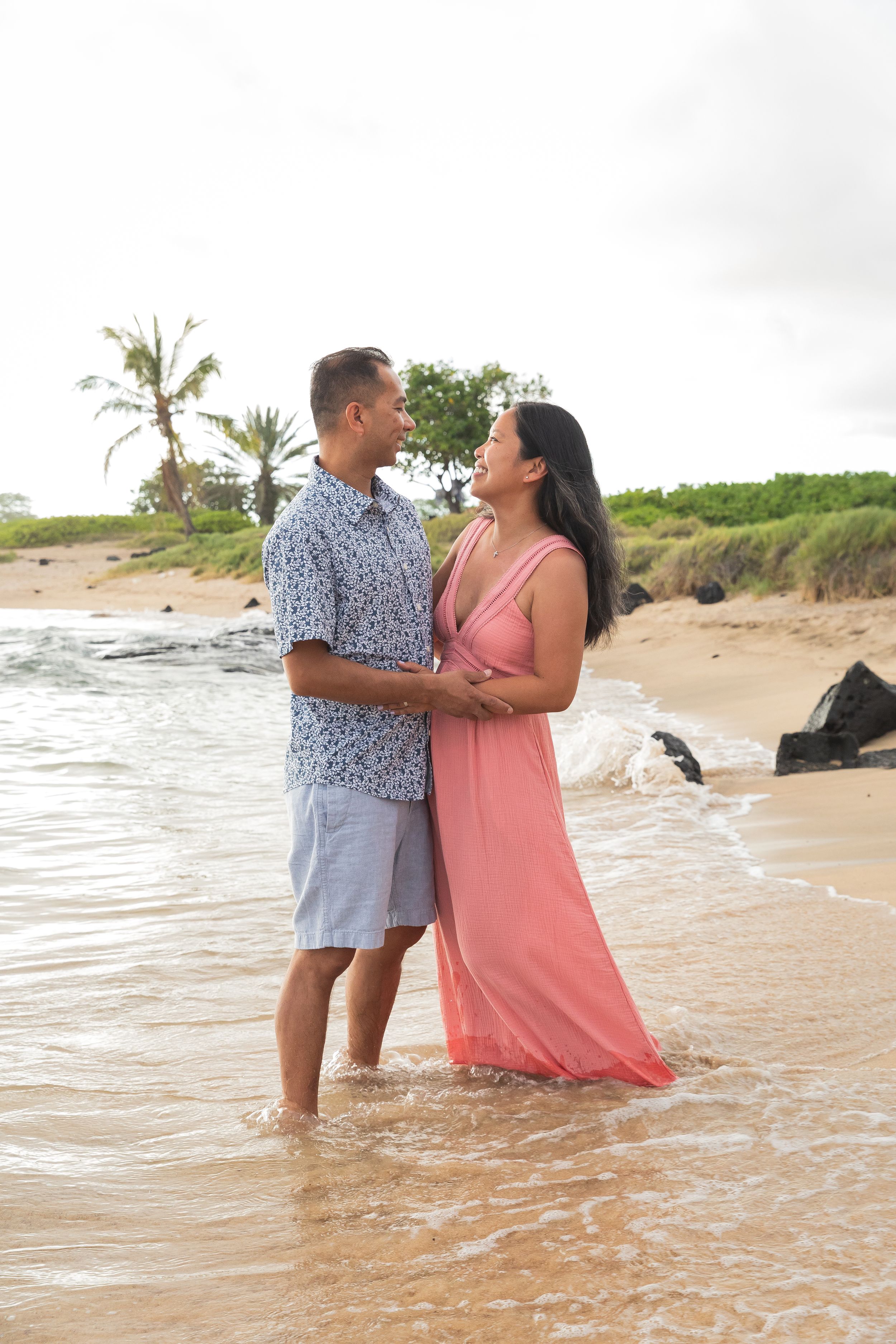 A couple stands in shallow ocean water on a tropical beach with palm trees in the background.