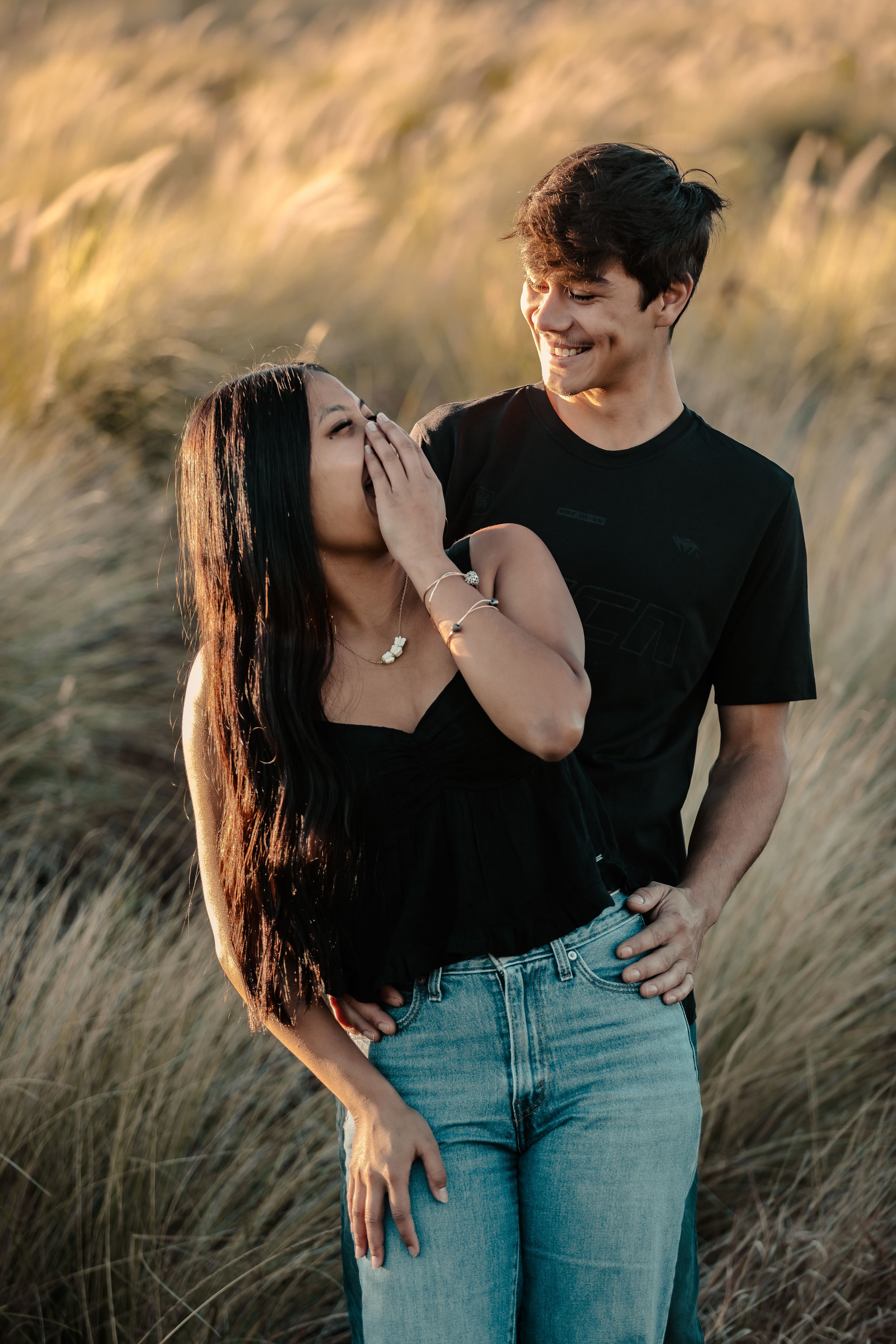 A couple shares a candid moment together in tall grass during golden hour.