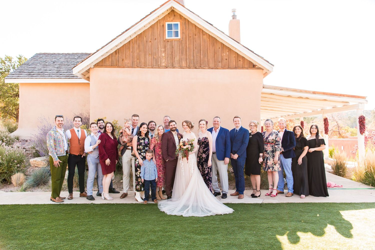 Large wedding party poses for a group photo outside a beige building with the desert landscape behind them.