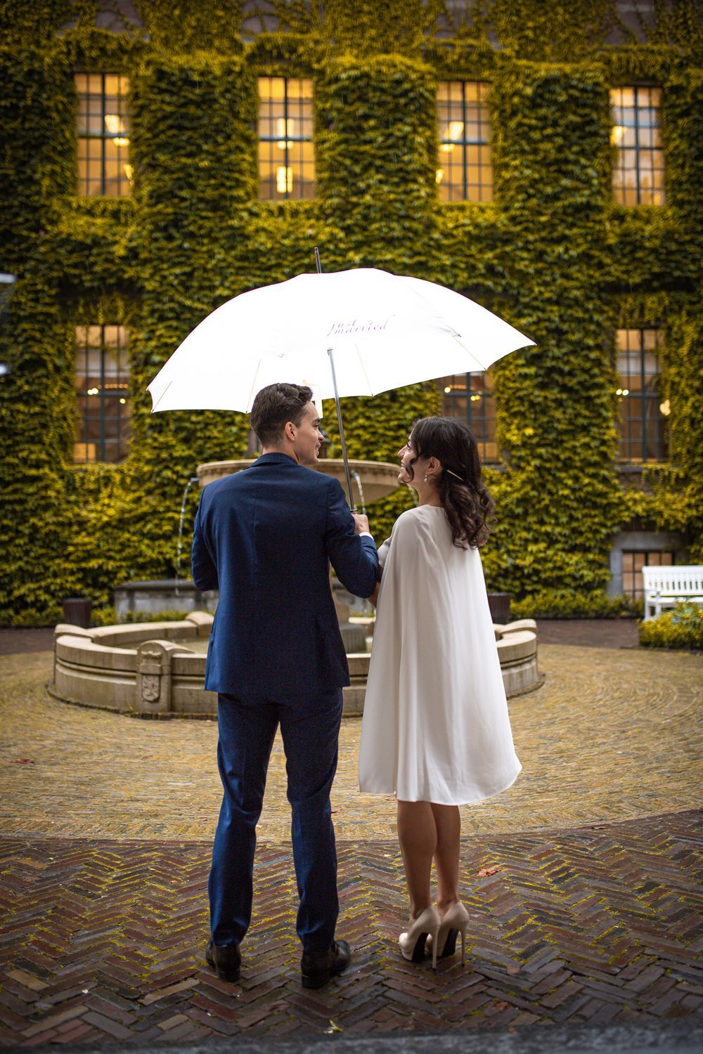 A couple shares a romantic moment under a white umbrella against an ivy-covered building wall after their marriage ceremony