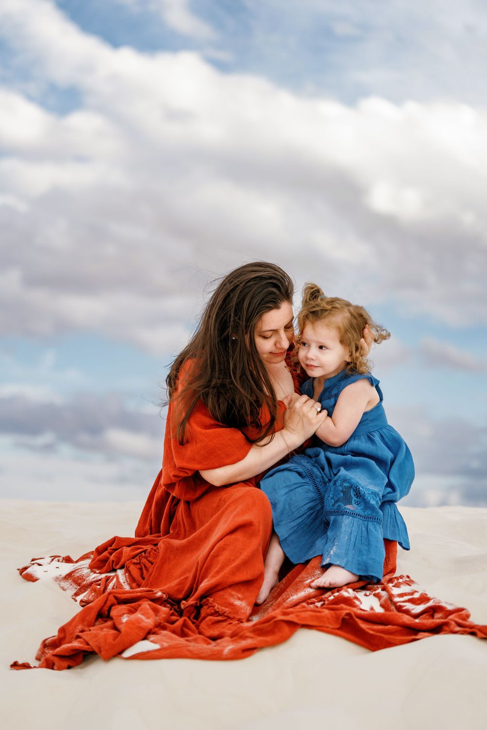 A tender moment shared between parent and child sitting on white sand dunes under cloudy skies.