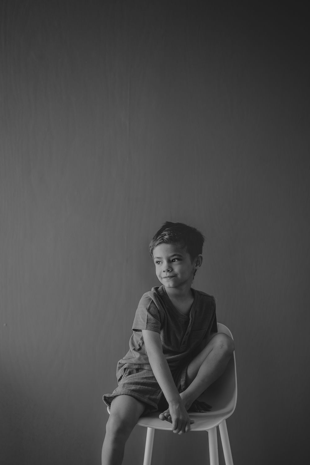 Artistic black and white portrait of child seated on white stool against minimal background.