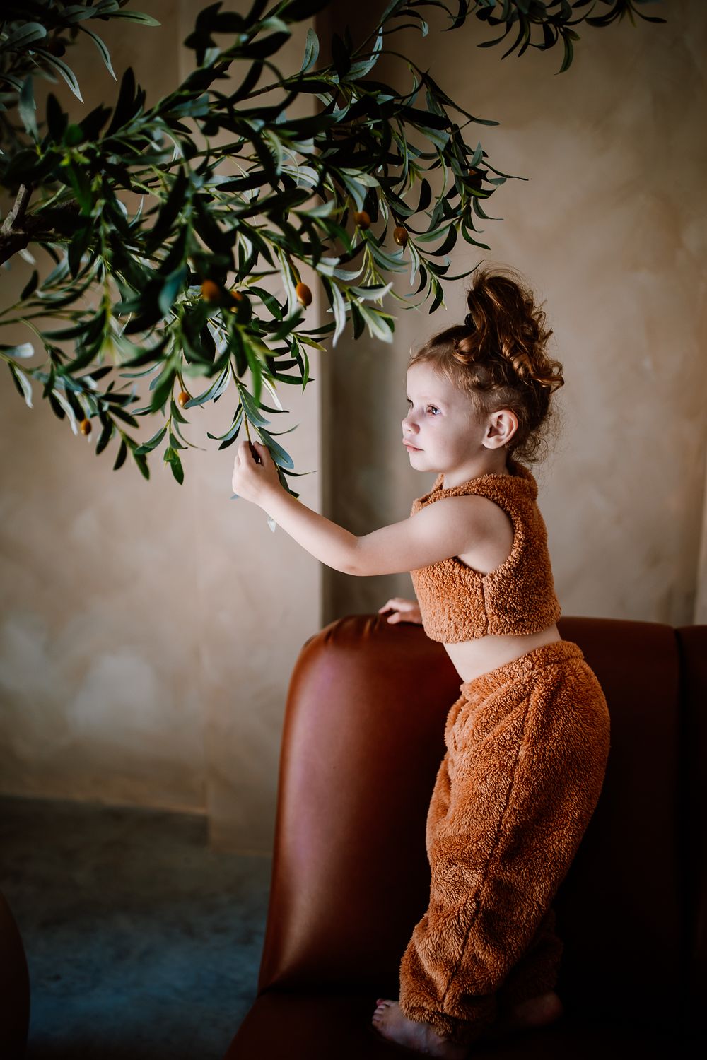 Small child in fuzzy orange outfit reaching up to touch olive branches.