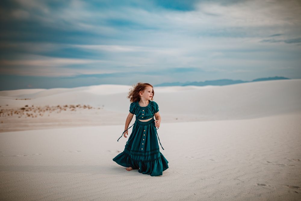 Figure in teal dress walking through white sand dunes under cloudy sky.