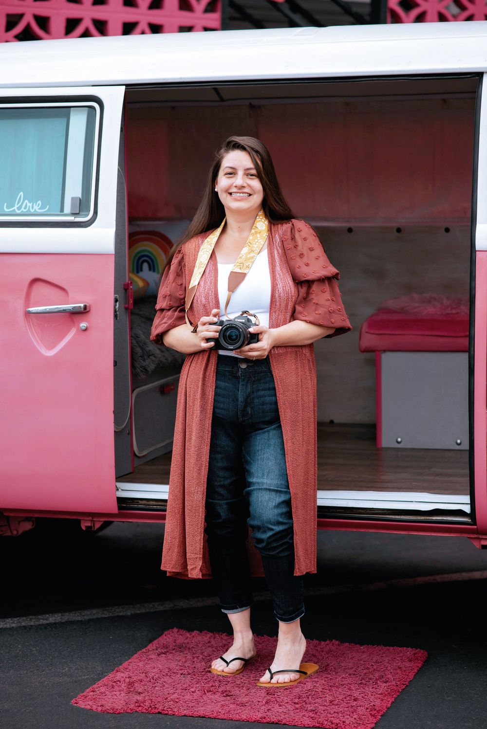 Photographer with vintage camera stands on pink rug beside retro pink VW van wearing rust-colored cardigan and jeans.