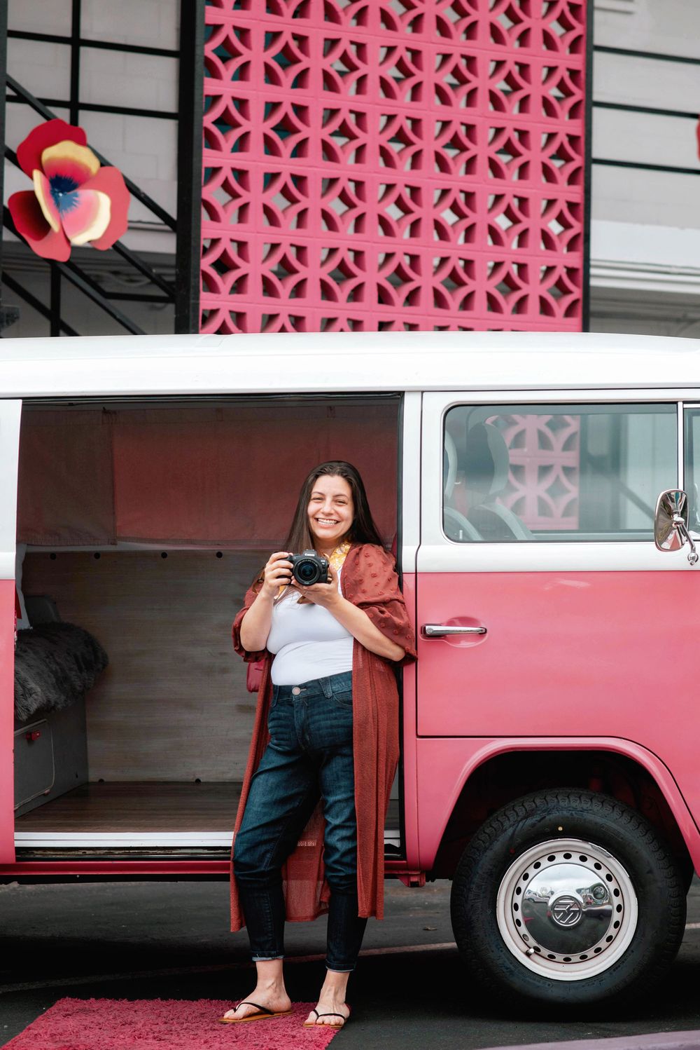 A candid lifestyle shot of someone holding a drink while standing next to a pink vintage VW van with a decorative wall.