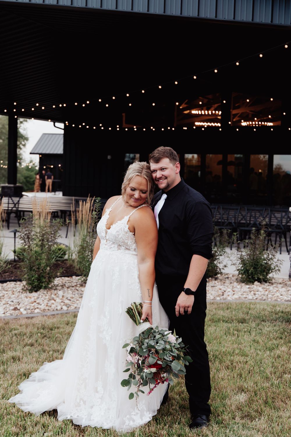 Romantic outdoor wedding portrait under string lights with rustic venue backdrop.