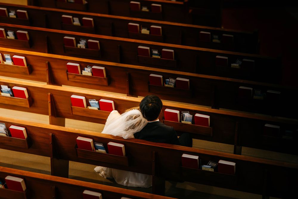Two people sit together in empty wooden church pews illuminated by warm lighting.
