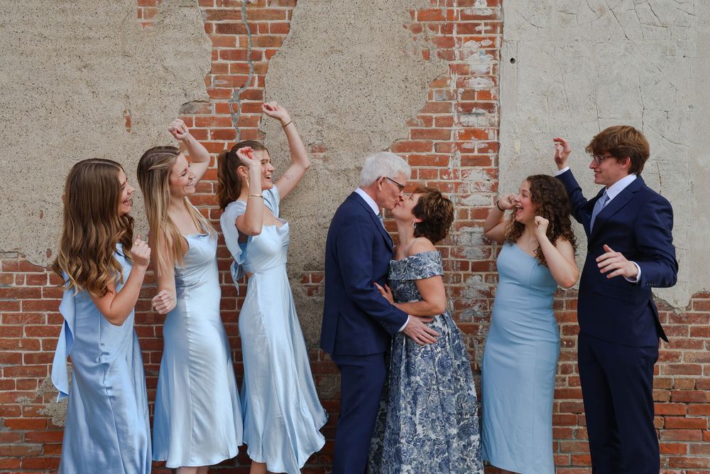 A wedding party in light blue dresses and navy suits celebrates joyfully against a rustic brick wall backdrop.