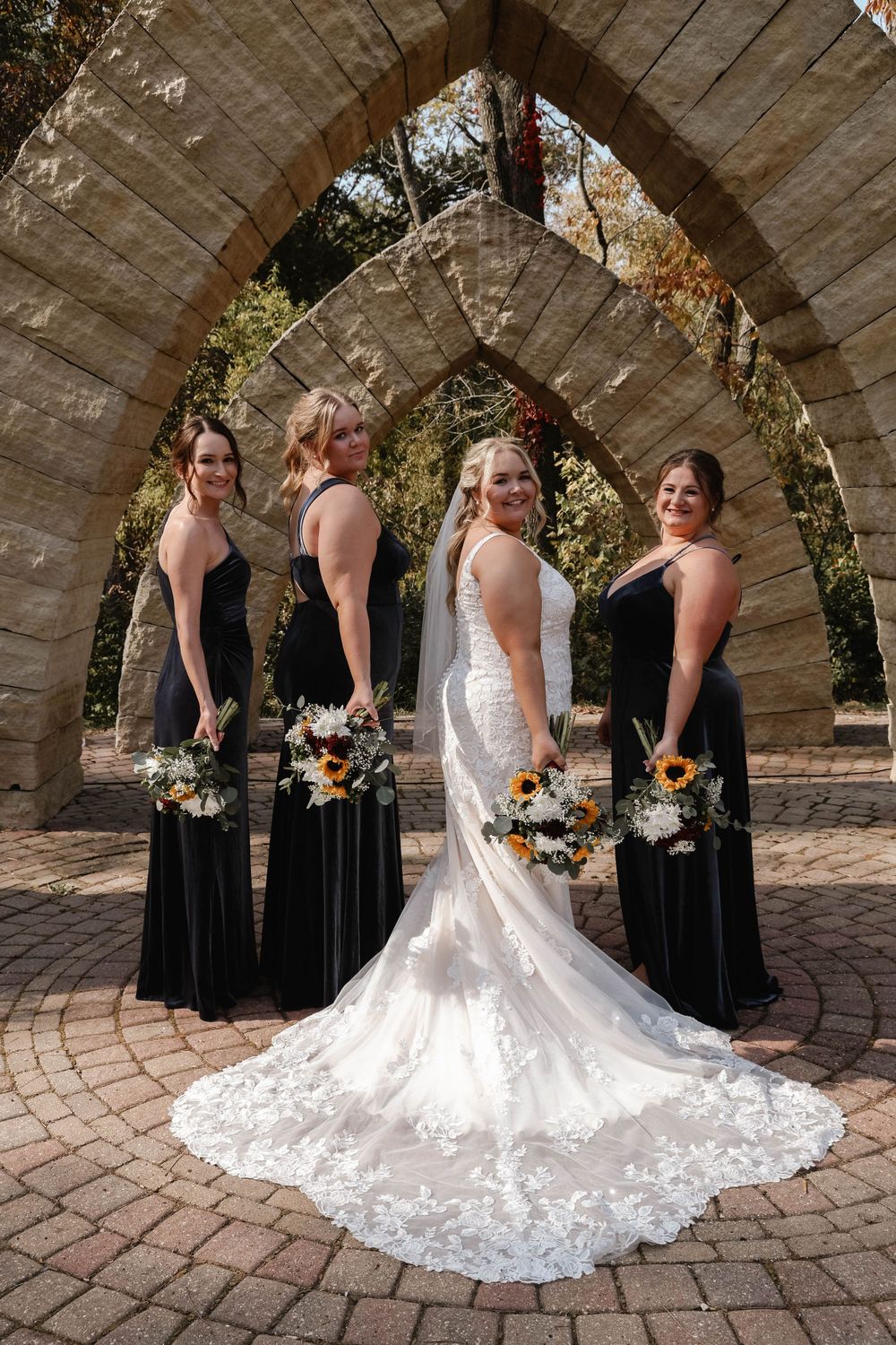 Bride in white lace gown with bridesmaids in black dresses under a stone archway.