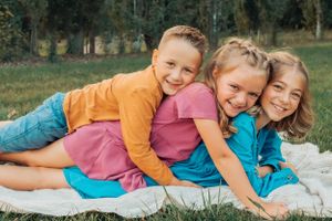 Three children in bright clothing cuddle together while lying on white blanket outdoors.