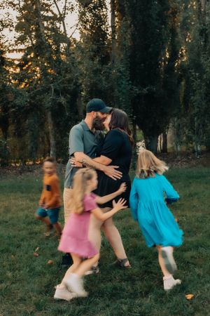 A candid outdoor family moment shows children in colorful dresses playing around embracing adults at sunset.