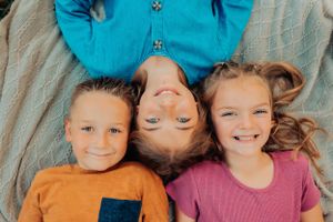 Three children lying down head-to-head on a blanket, wearing bright colored clothing.