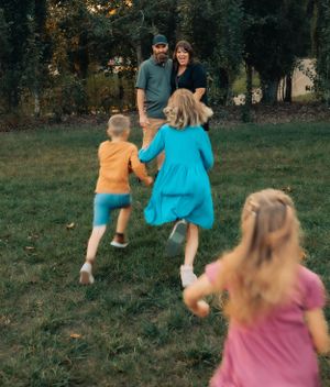 A playful scene of three children running towards two adults in a park setting at sunset.