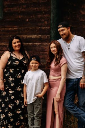 A casual family portrait against a rustic wooden wall background shows coordinated outfits and warm smiles.