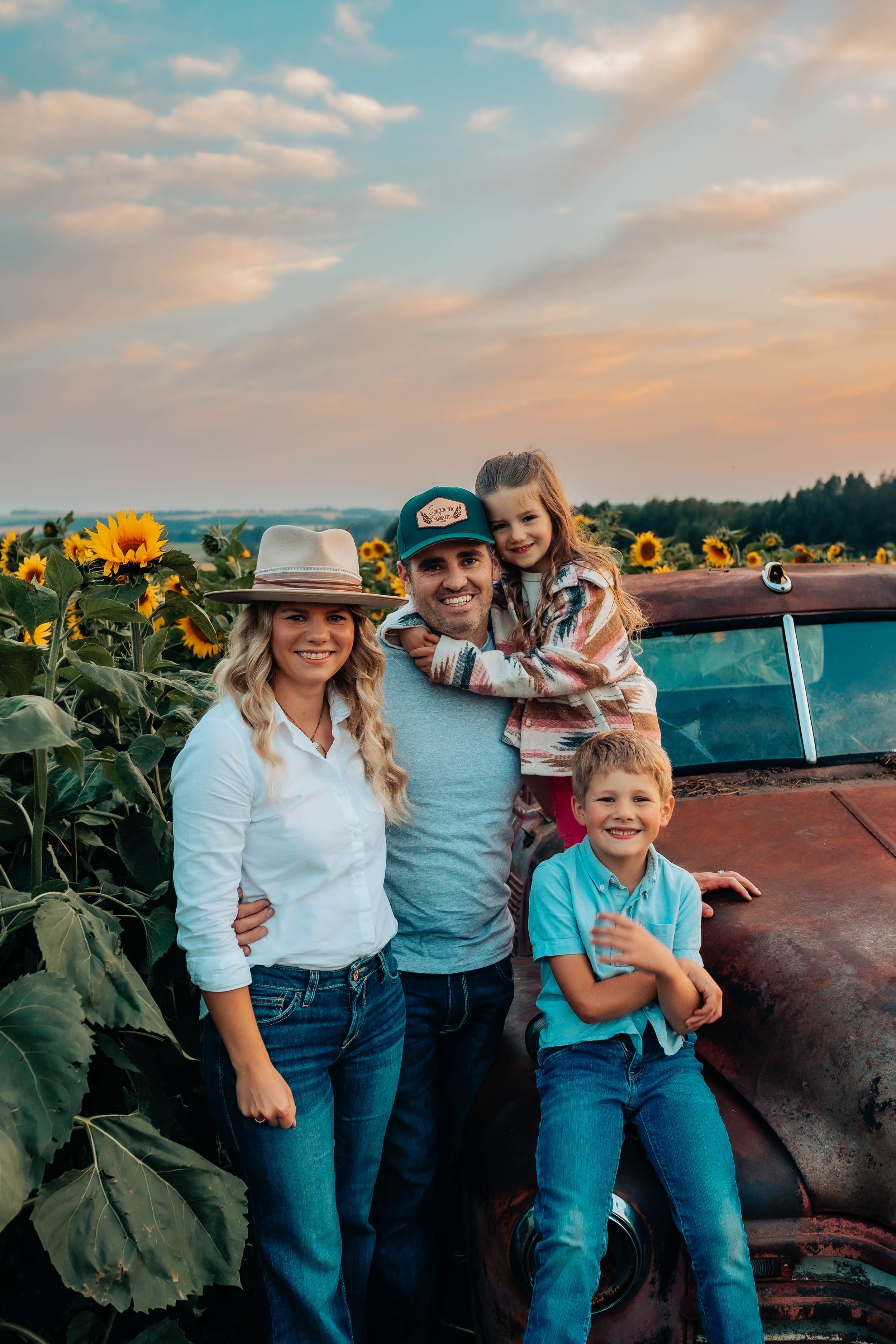 Family poses near vintage truck in sunflower field during golden hour.