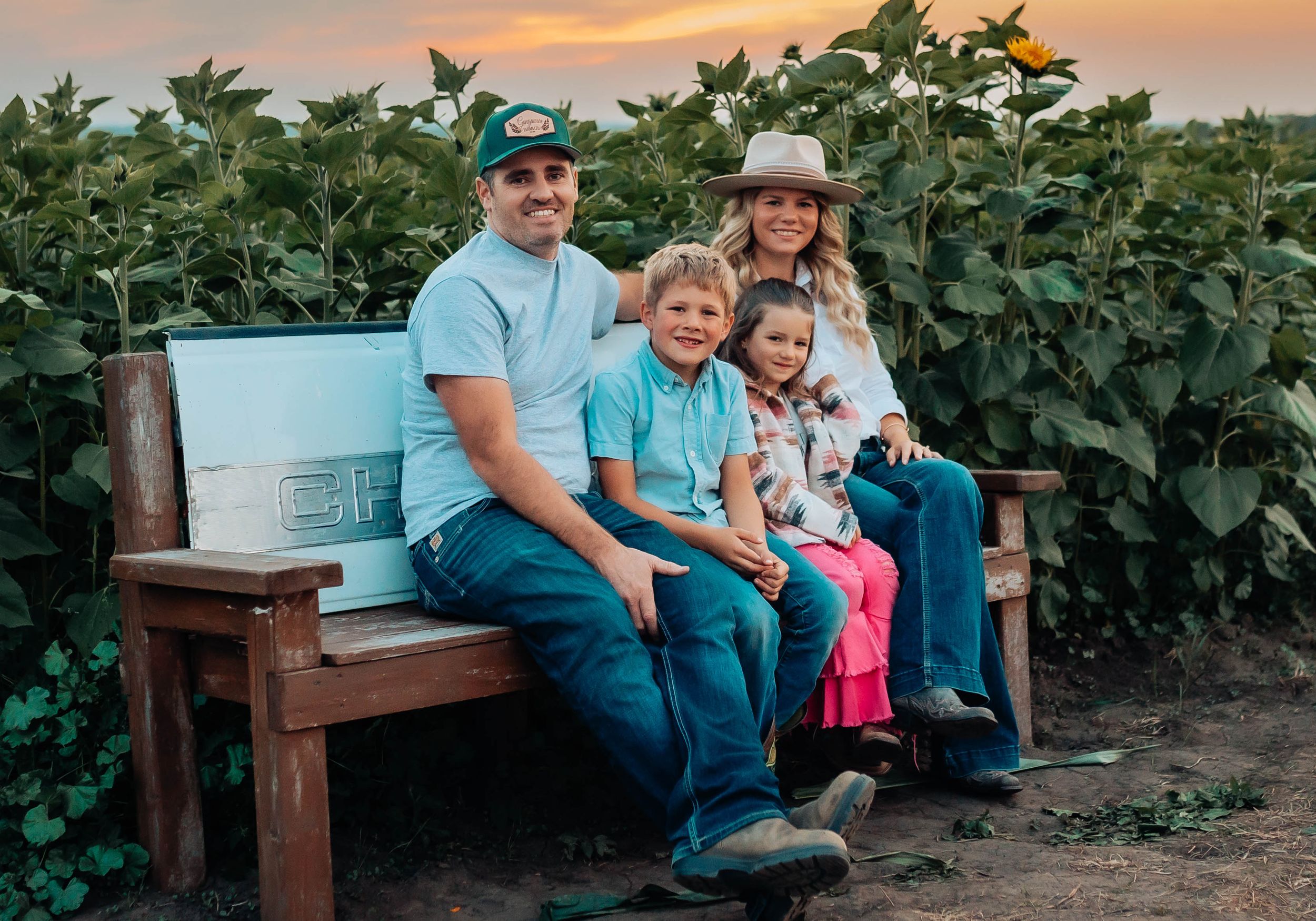 Family sits together on wooden bench at sunset in front of lush greenery.