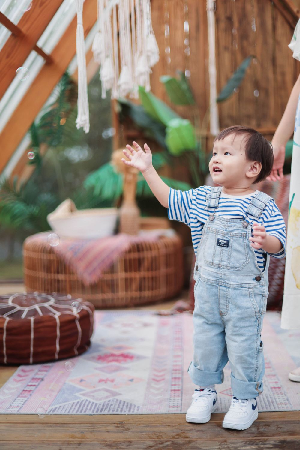 Happy toddler in denim overalls reaching up toward hanging decorations in boho interior.