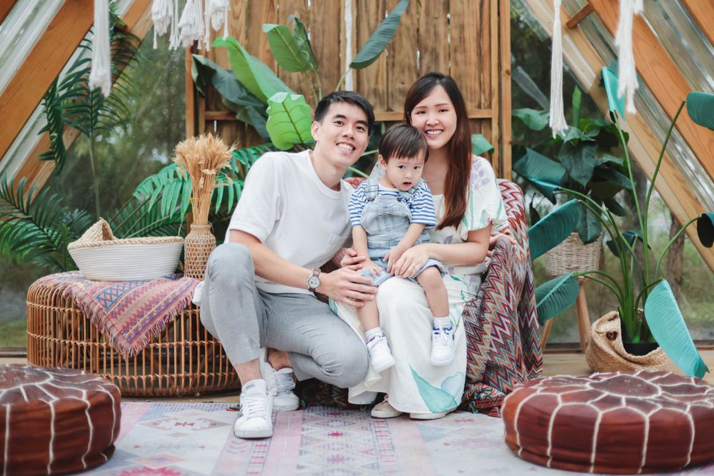 Asian family of three sitting together smiling in a tropical boho-decorated space.