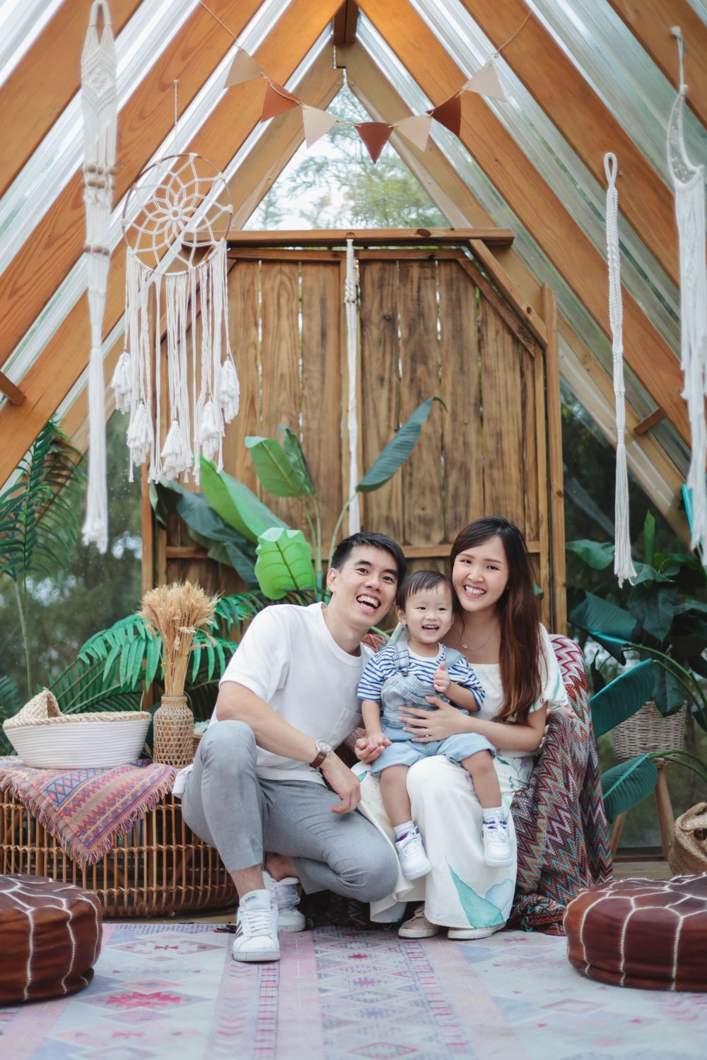 Happy Asian family with toddler posing inside a boho-styled wooden cabin with plants.