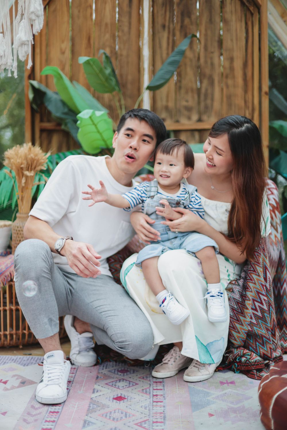 Asian family with young toddler posing together in a boho-styled outdoor setting with tropical plants.