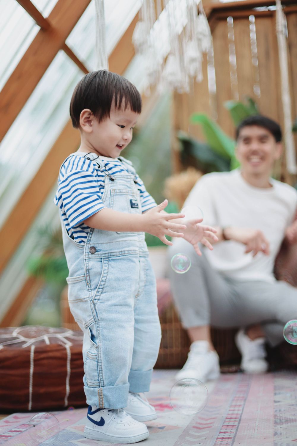 Toddler boy in denim overalls playing with bubbles while father watches in boho decorated space.