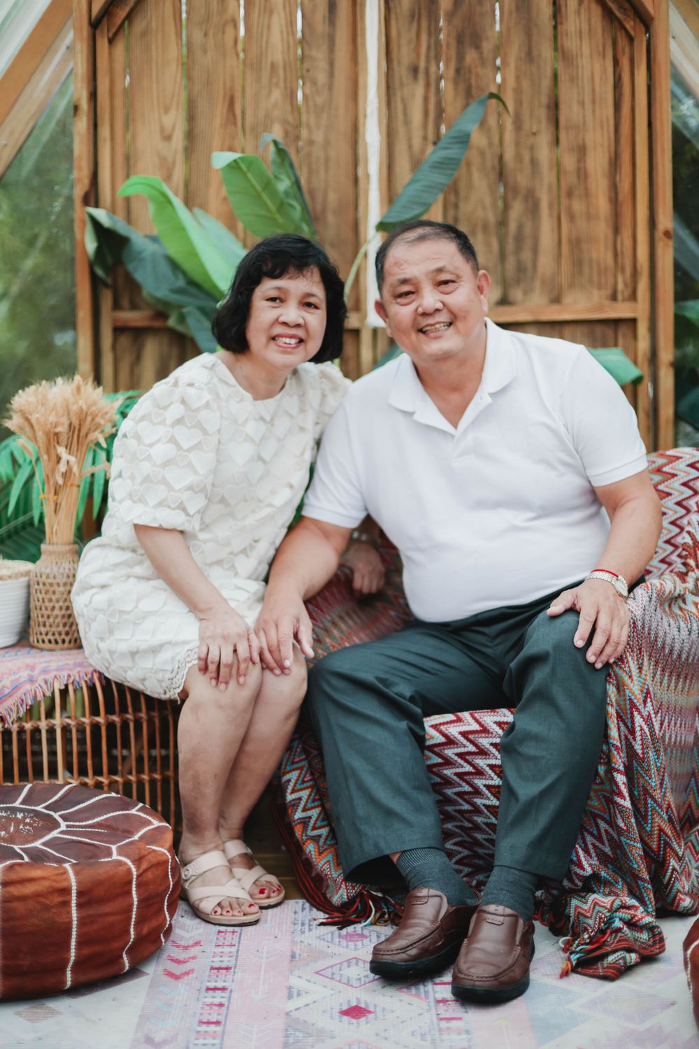 Smiling Asian senior couple sitting together on colorful cushions in a tropical decorated outdoor space.
