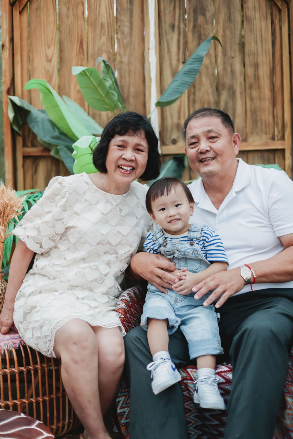 Happy Asian grandparents sitting with adorable toddler grandchild in a tropical outdoor setting.