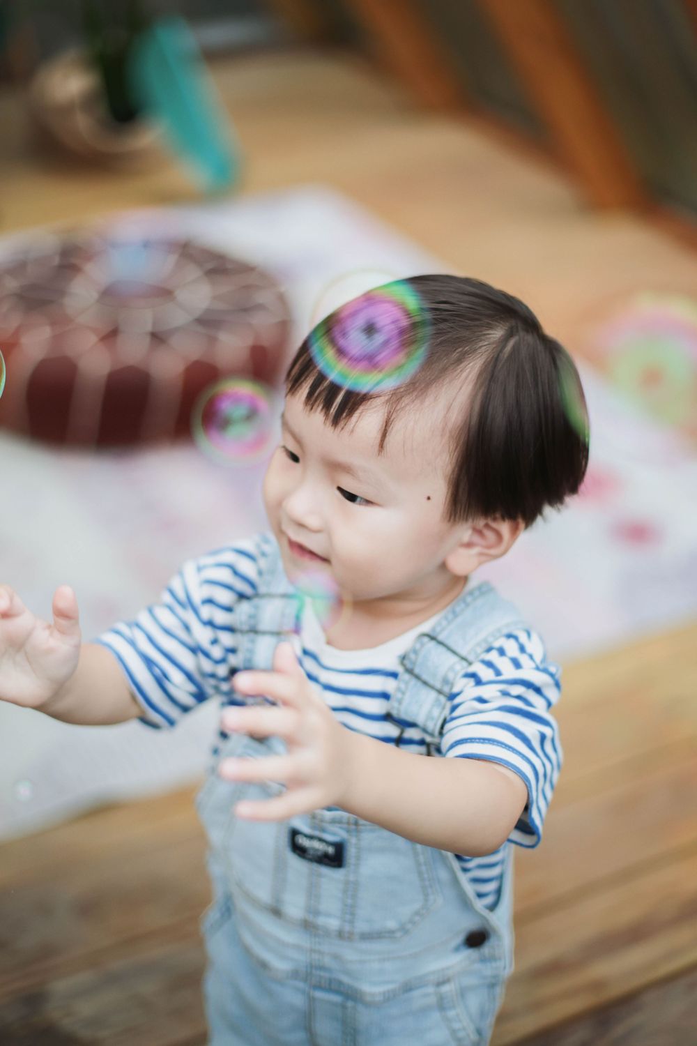 Cute Asian toddler boy in striped shirt and overalls reaching for colorful soap bubbles outdoors.