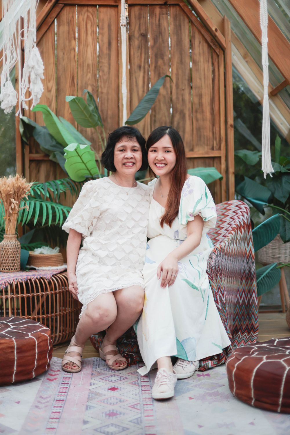 Two Asian women, mother and daughter, smiling together in a boho-styled tropical outdoor setting.